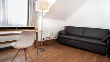 A modern living room with a wooden floor, a black leather sofa, a white chair, a small table with a lamp, and a window with sheer curtains.