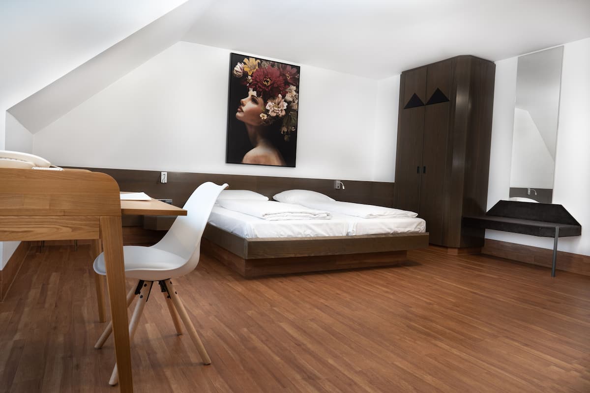 A bedroom with a wooden floor, a bed with white bedding, a desk with a chair, and a large painting on the wall.