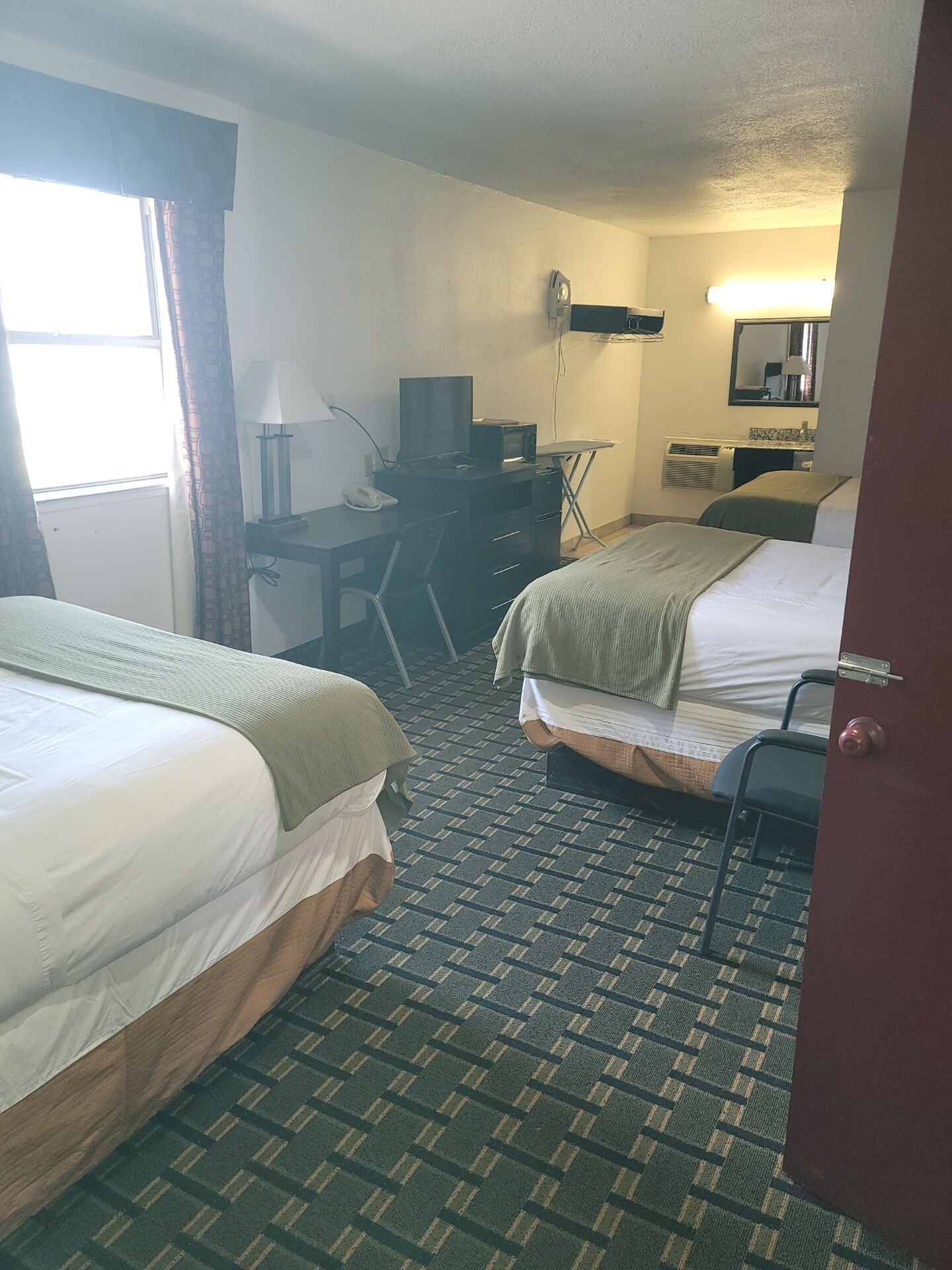 A hotel room with two beds, a desk with a computer, a chair, a desk lamp, a mirror, and a window with curtains.