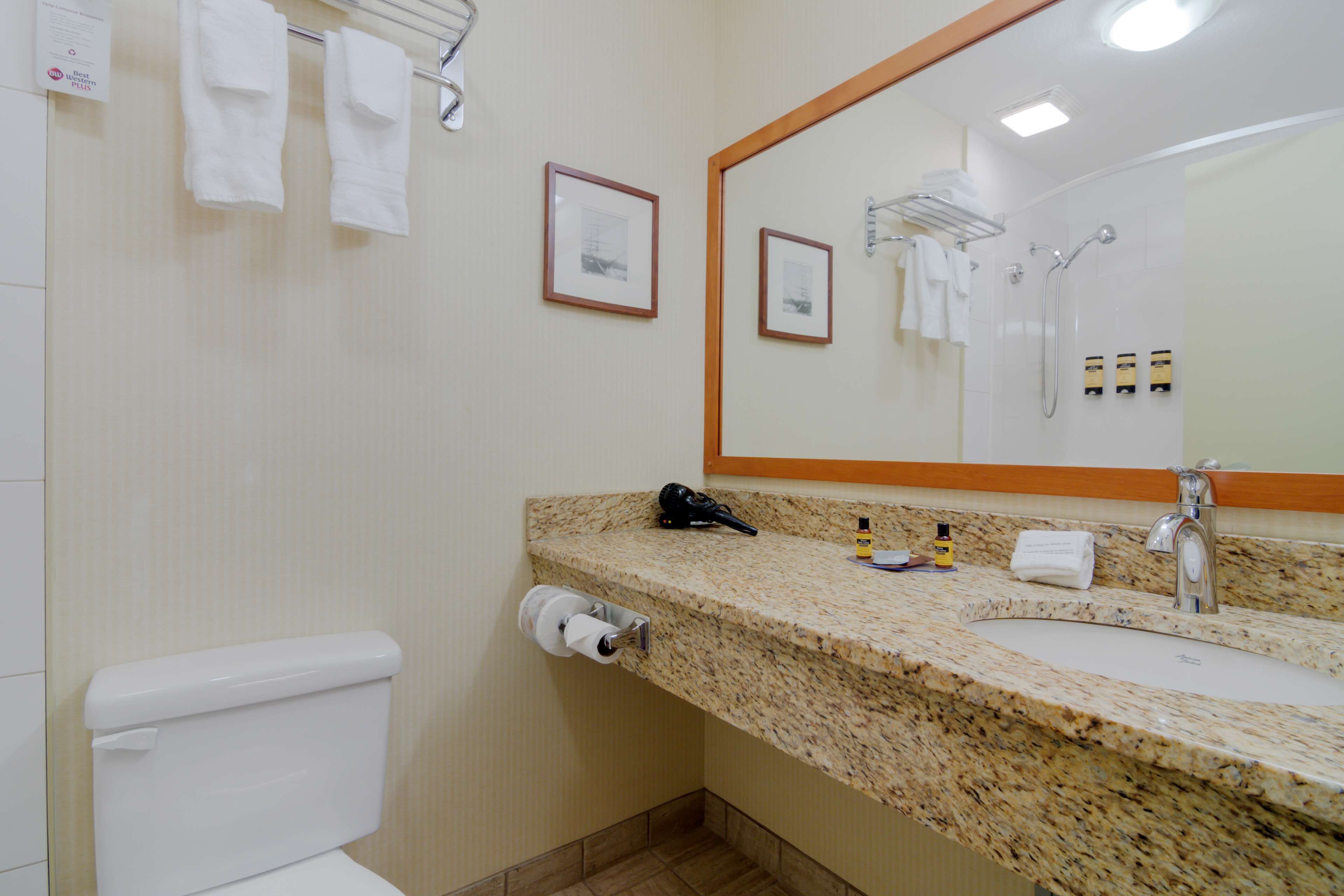 A bathroom with a toilet, sink, and mirror. There are towels hanging on a rack and a framed picture on the wall.