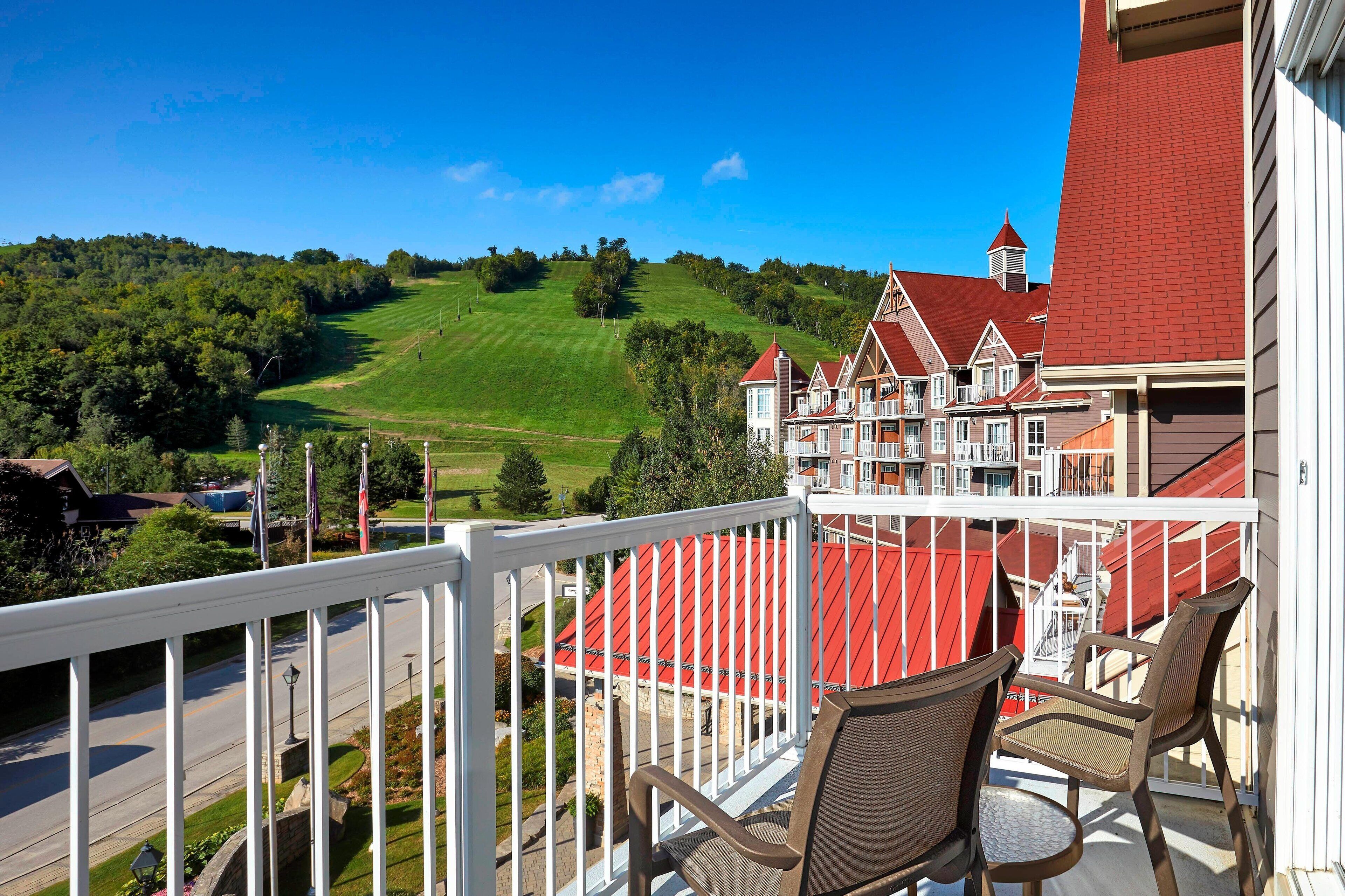A balcony with a view of a red-roofed building, a grassy hill, and a road.