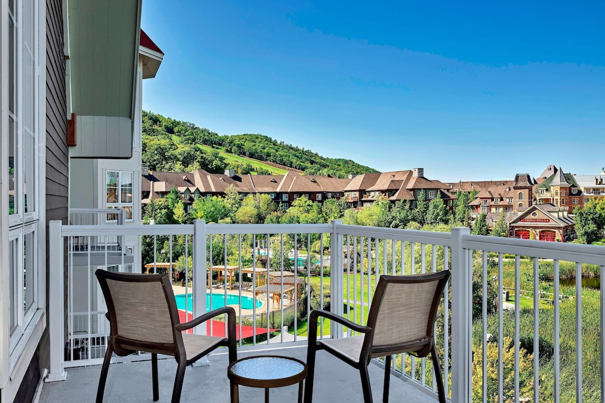 A balcony with two chairs and a small table, overlooking a pool and a landscaped area with buildings in the background.