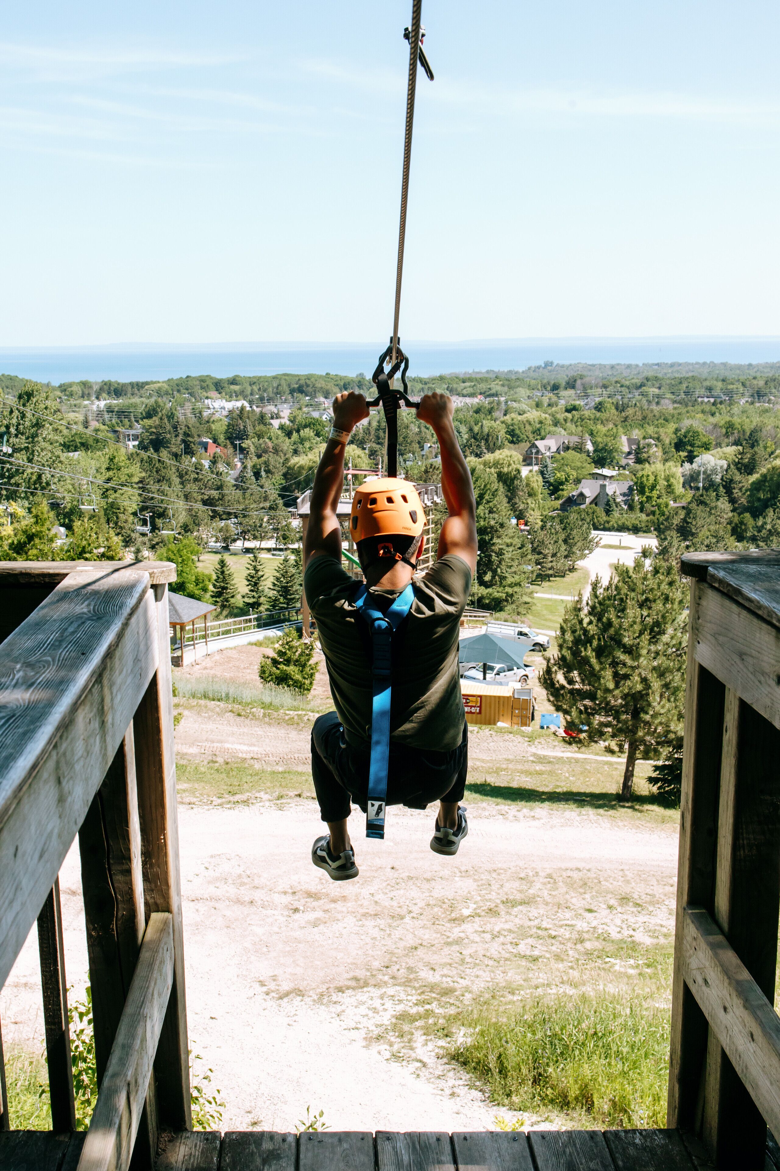A person in a helmet and harness suspended by a rope, with a view of a wooded area and buildings in the background.