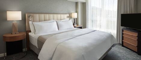 A hotel room with a large bed, two bedside tables with lamps, a flat-screen TV, and a large window with curtains.