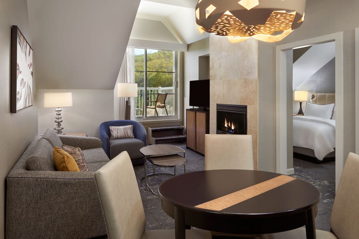 A modern hotel room with a sofa, armchairs, a round table, a fireplace, and a balcony with a view of the outdoors.