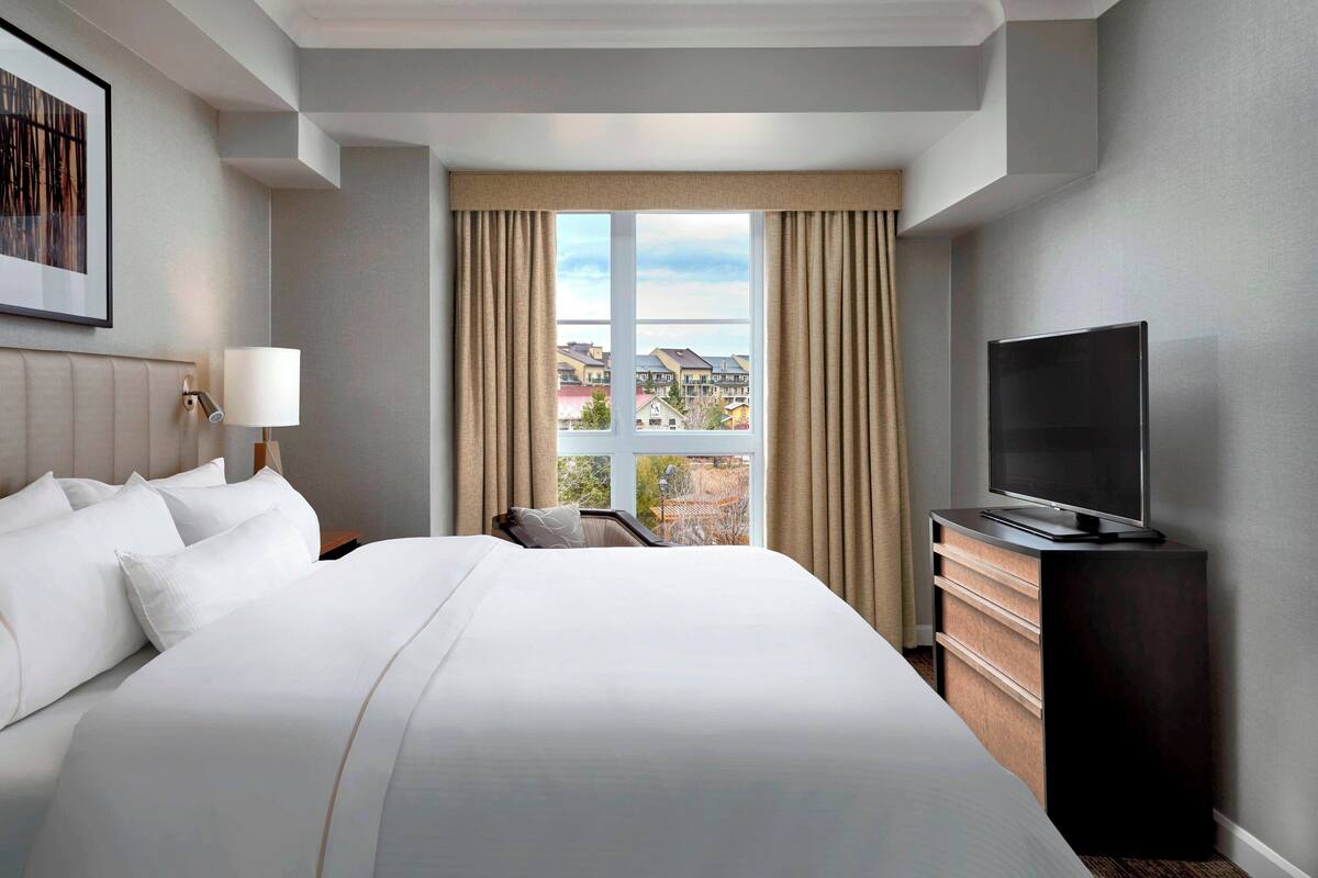 A hotel room with a large bed, a flat-screen TV, a nightstand with a lamp, and a view of buildings through the window.