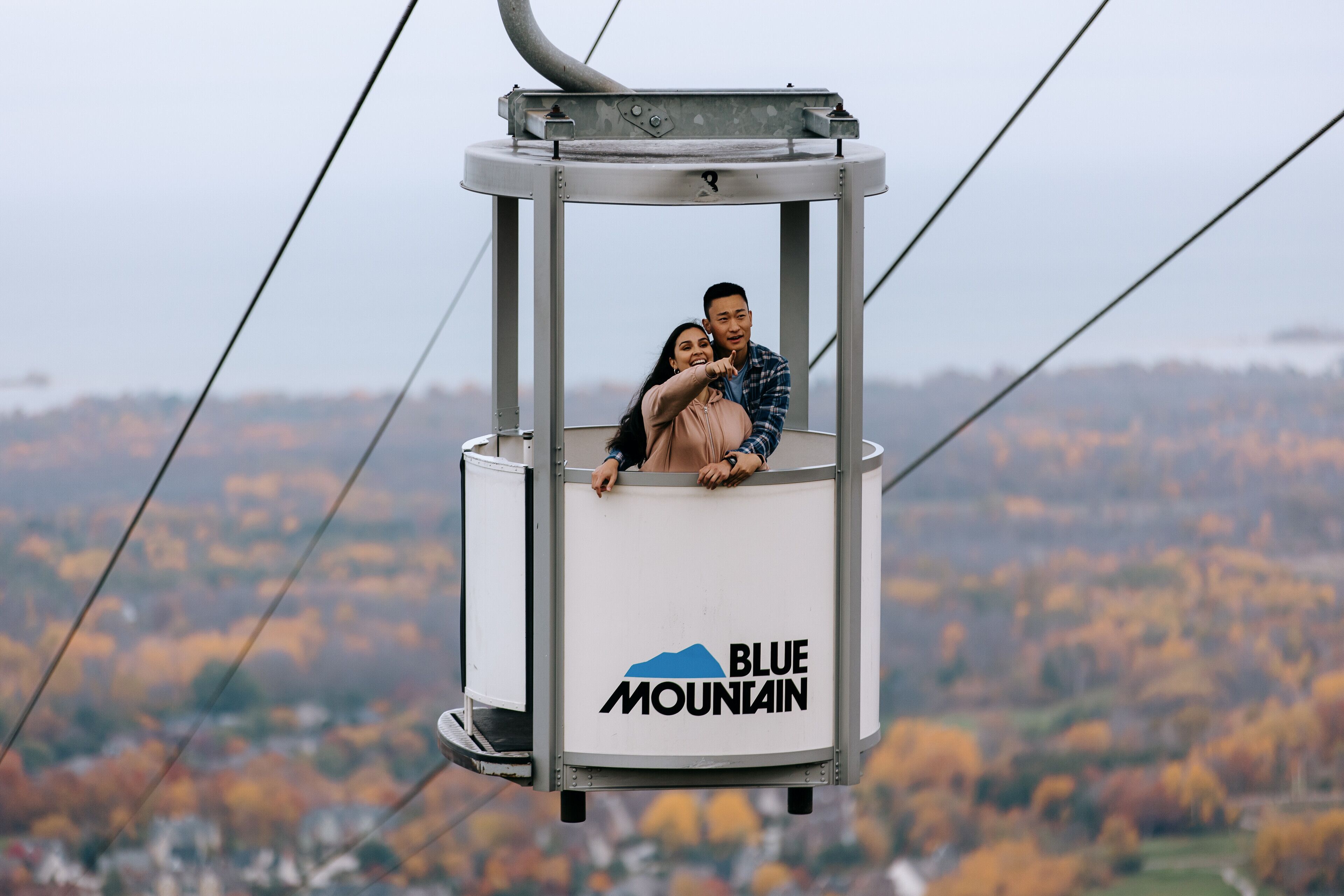 A cable car with a logo labeled "Blue Mountain" suspended above a landscape with autumn-colored trees.