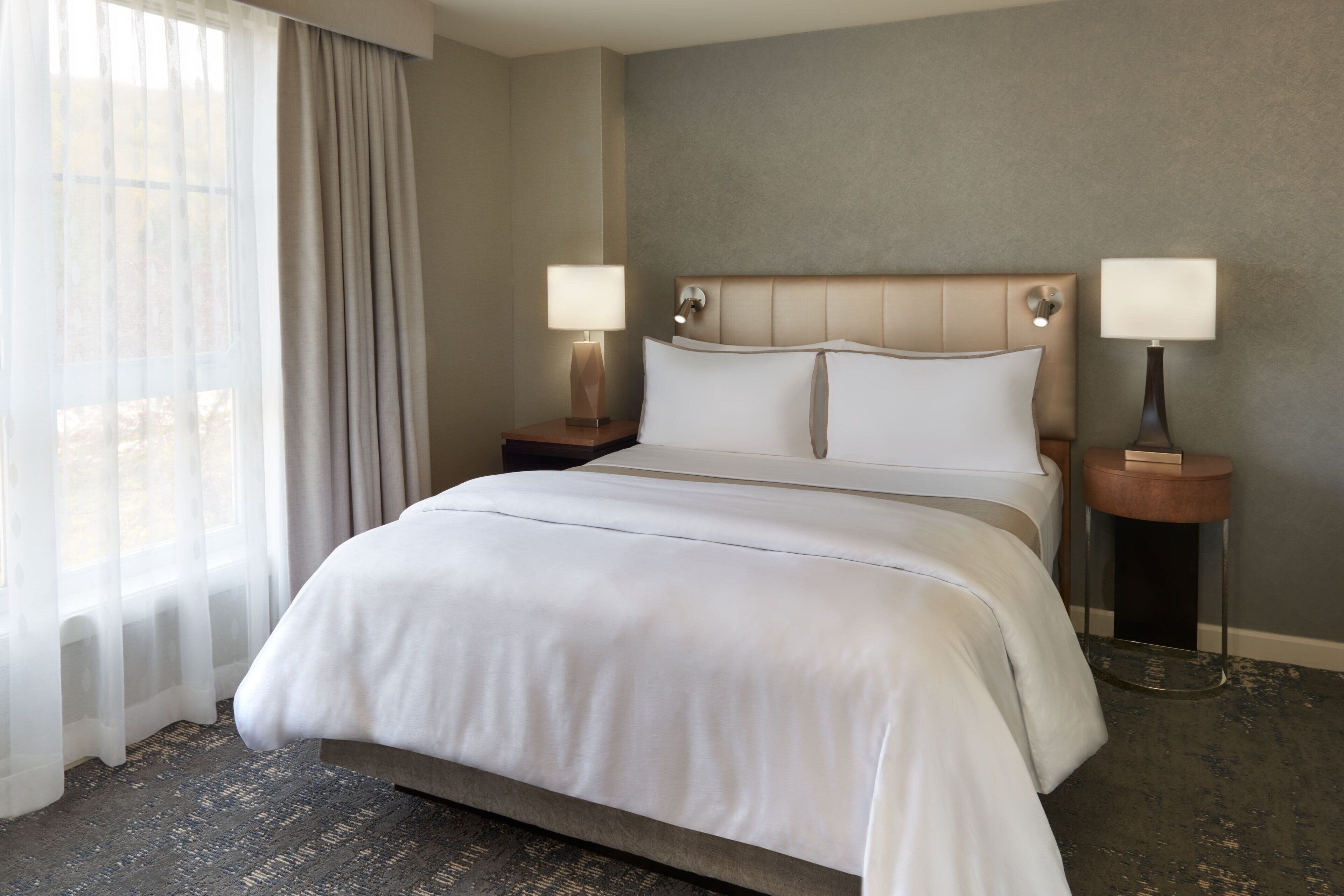 A hotel room with a large bed, two bedside tables with lamps, a window with sheer curtains, and a textured carpet.