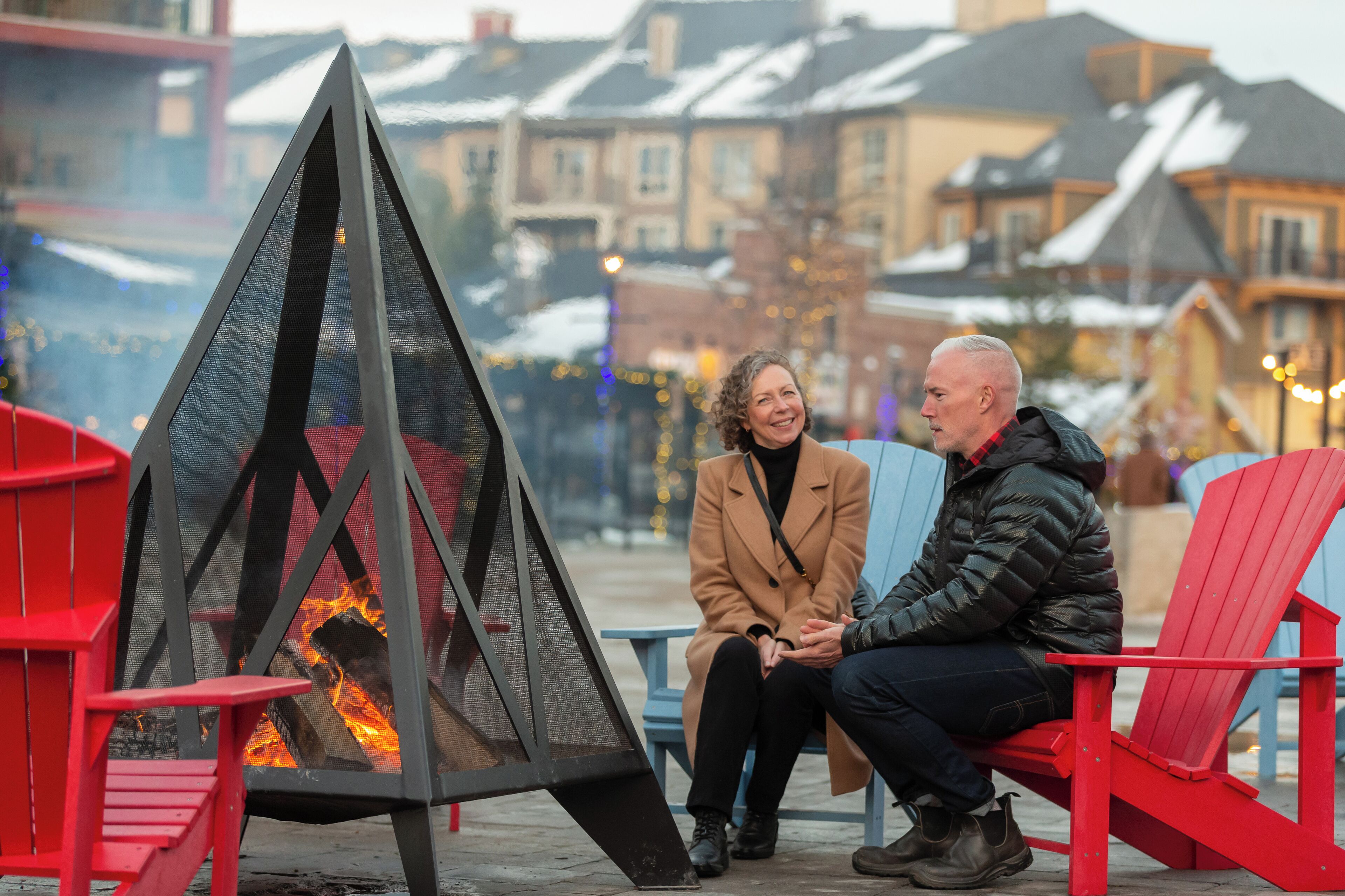 Two people seated on red chairs near a fire pit with a view of snow-covered buildings in the background.