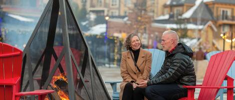 Two people seated on red chairs near a fire pit with a view of snow-covered buildings in the background.