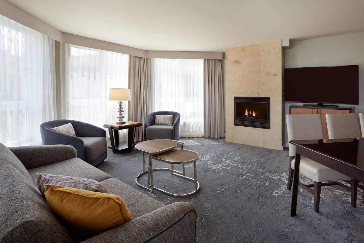 A modern hotel room with a grey sofa, a round coffee table, a TV, a fireplace, and a dining area with a table and chairs.