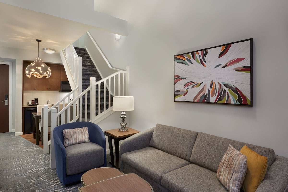 A modern living room with a gray sofa, a blue armchair, and a wooden coffee table. A staircase with a white railing leads to a kitchen area.