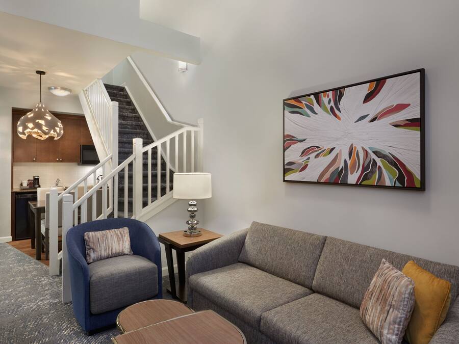 A modern living room with a gray sofa, a blue armchair, and a wooden coffee table. A staircase with a white railing leads to a kitchen area.