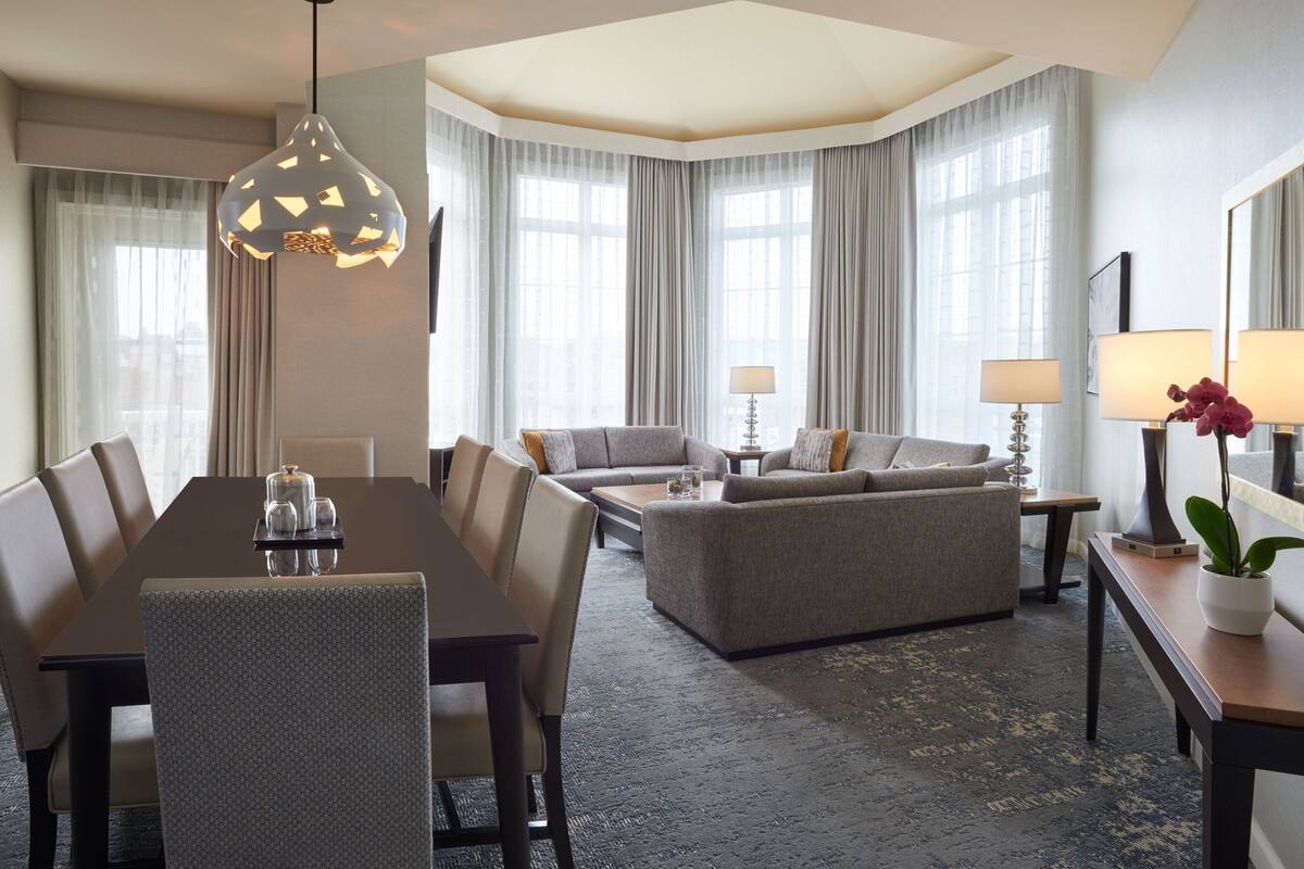 A hotel room with a dining table, sofa, and a large window with curtains.