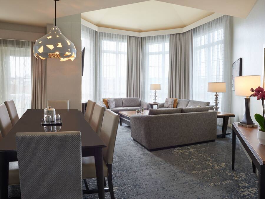 Habitación de hotel con mesa de comedor, sofá y un ventanal con cortinas.