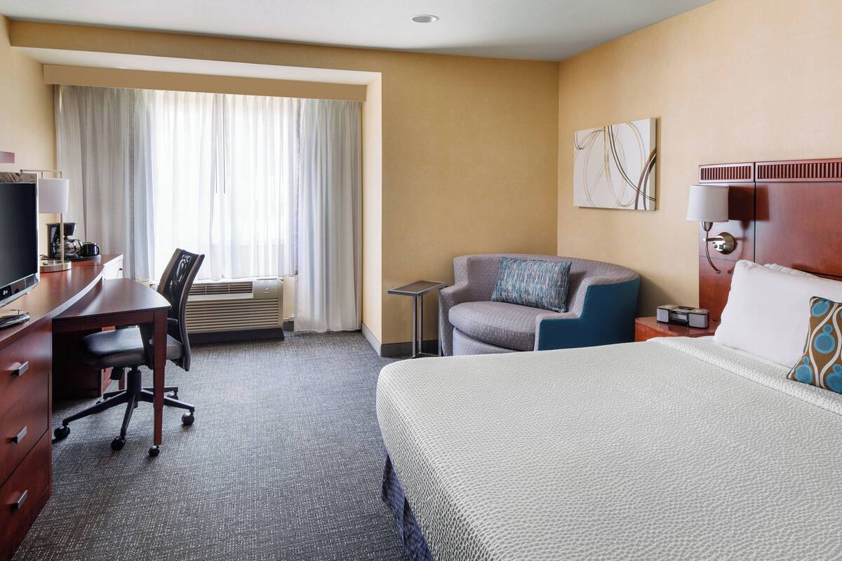 Een hotelkamer met een bed, een bureau met stoel, een klein tafeltje, een televisie en een raam met gordijnen.