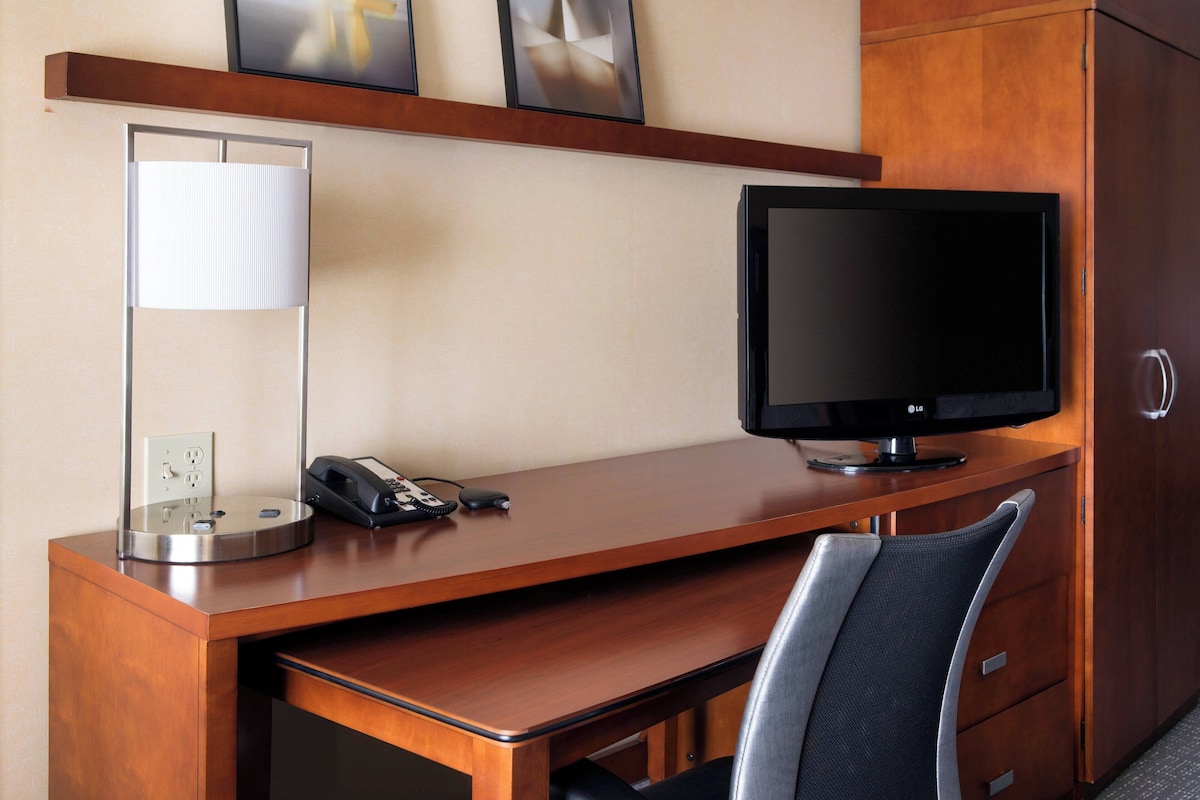 A hotel room with a wooden desk, a flat-screen TV, a telephone, and a lamp.