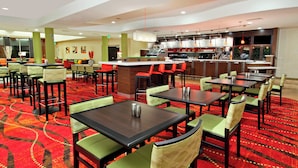 En hotelrestaurant med flere spiseborde og stole, et barområde og et køkken i baggrunden.