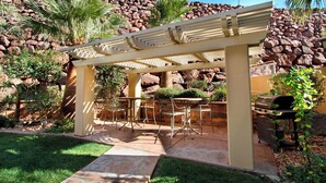 En udendørs terrasse med pergola, siddepladser og grill.