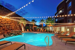 A swimming pool with lounge chairs and string lights, surrounded by a stone wall and palm trees. - Courtyard by Marriott St George (St. George)