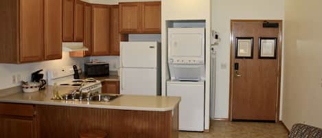 A compact kitchen with wooden cabinets, a microwave, a refrigerator, and a washing machine. There are two stools and a patterned chair.