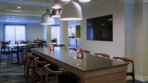 A hotel dining area with a long table, multiple chairs, pendant lights, and a flat-screen TV on the wall.