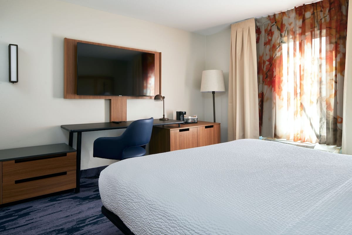 Una habitación de hotel con una cama grande, un escritorio con silla, un televisor de pantalla plana, una lámpara y un ventanal con cortinas.