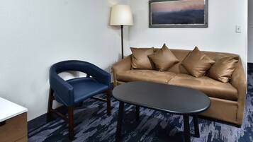 Una habitación con un sofá de cuero marrón, un sillón azul, una mesita redonda pequeña, una lámpara de pie y un cuadro en la pared.