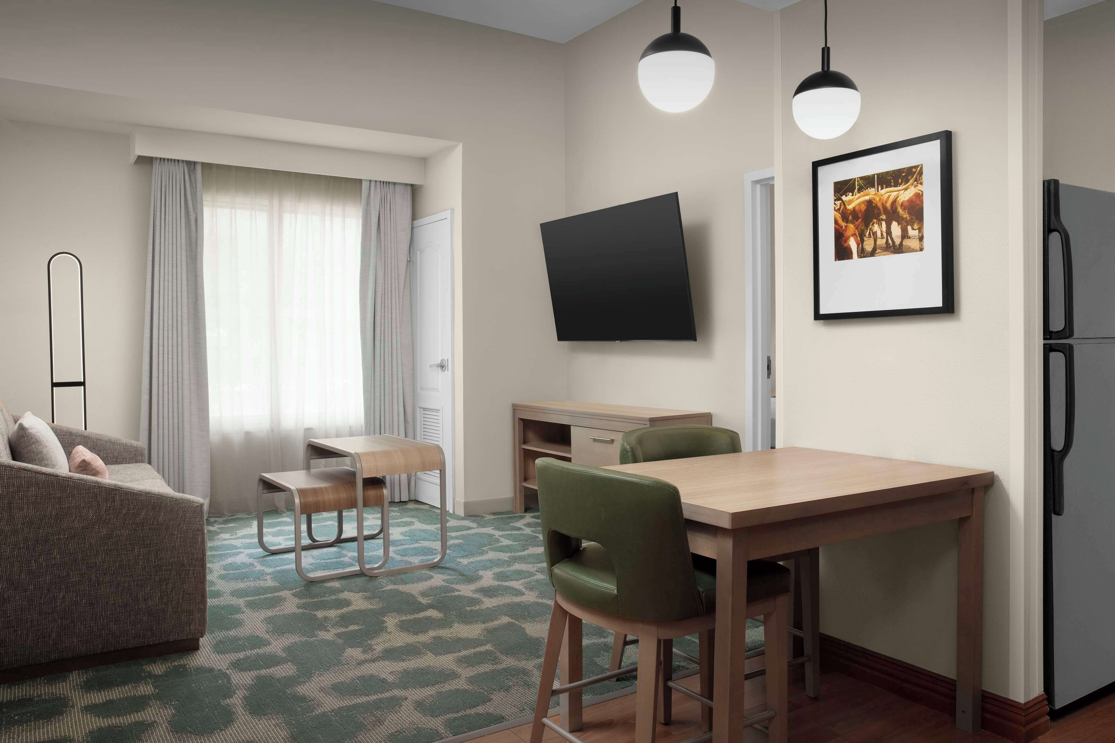 A modern hotel room with a sofa, a dining table with chairs, a flat-screen TV mounted on the wall, and a refrigerator.
