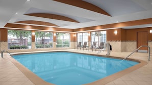 Une piscine intérieure à l’eau d’un bleu limpide, entourée d’un sol et de murs recouverts de carreaux beiges, avec quelques chaises et une porte visibles.