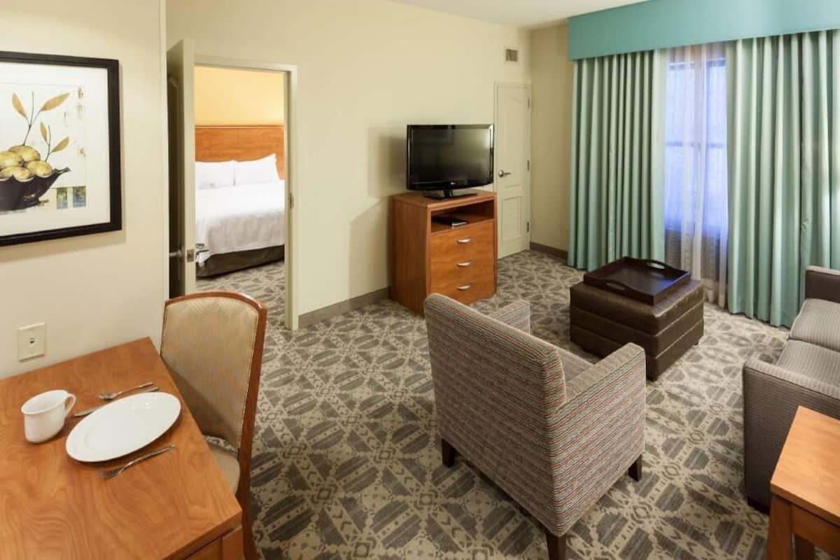 Une chambre d’hôtel avec un lit, un téléviseur posé sur un meuble en bois, une table à manger avec des chaises et une moquette à motifs.