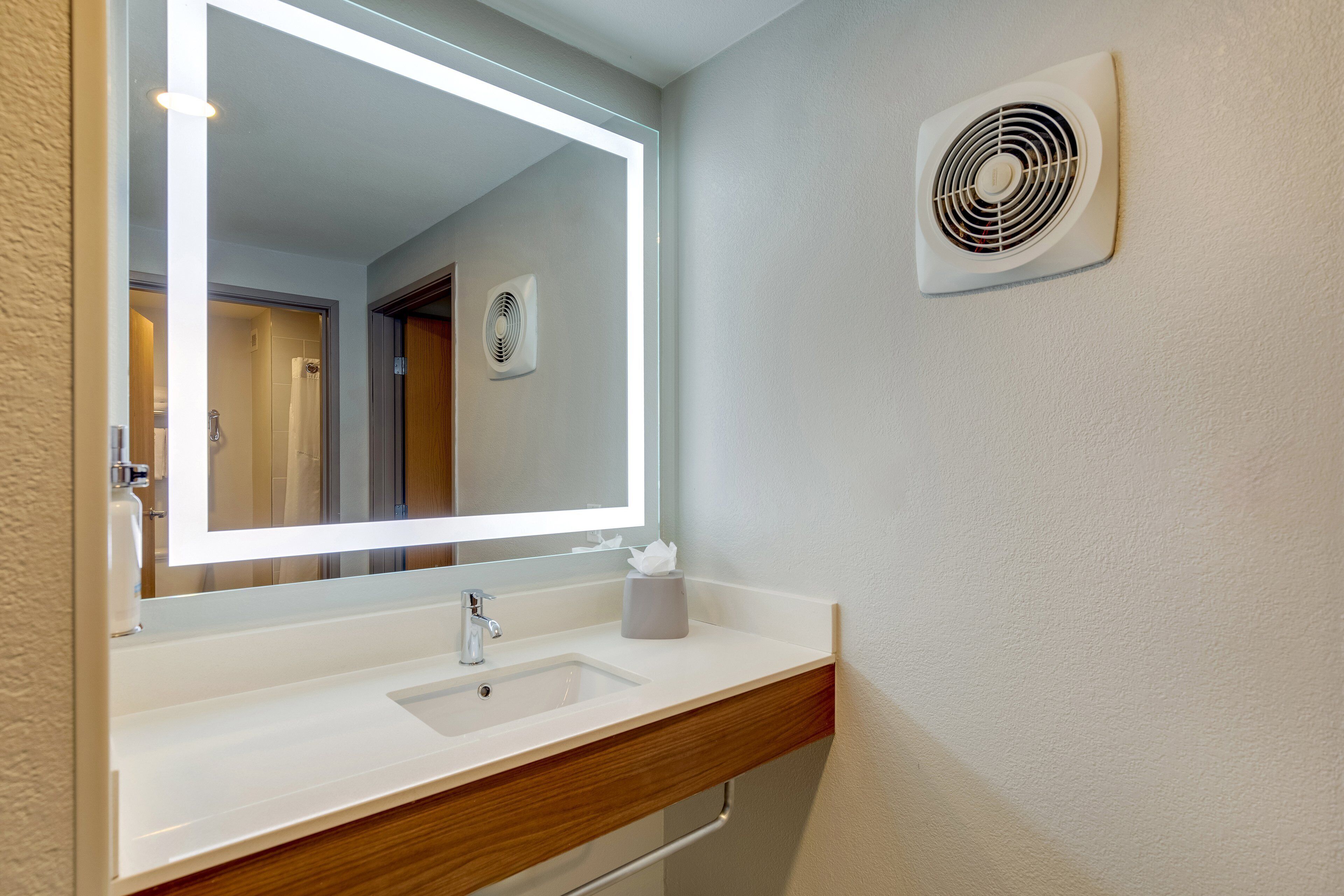 Un baño con un lavabo blanco, un espejo con iluminación integrada, un dispensador de toallas y un ventilador de pared.