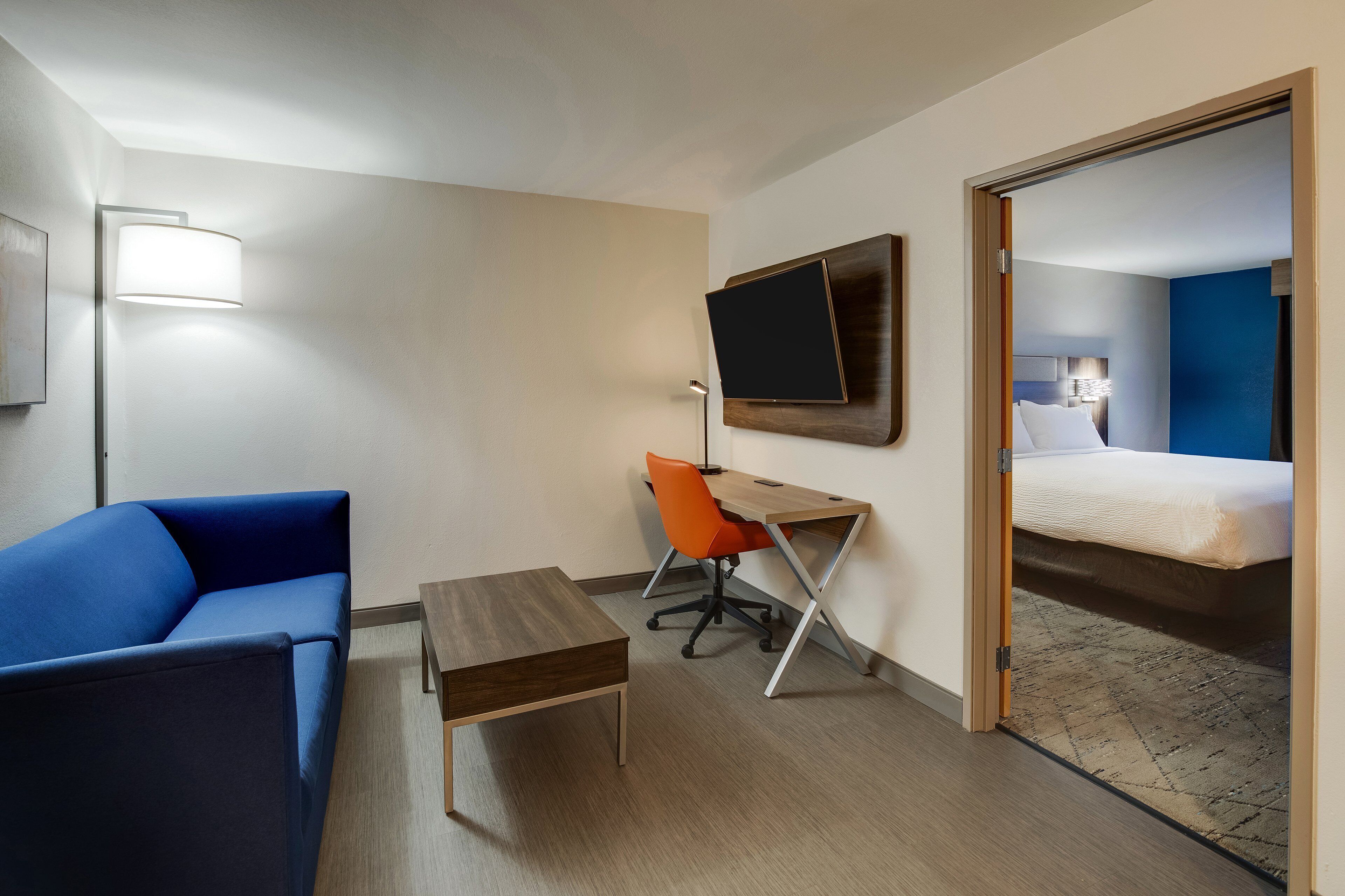 A hotel room with a blue sofa, a wooden desk, an orange office chair, a flat-screen TV mounted on the wall, and a bed with a blue headboard.