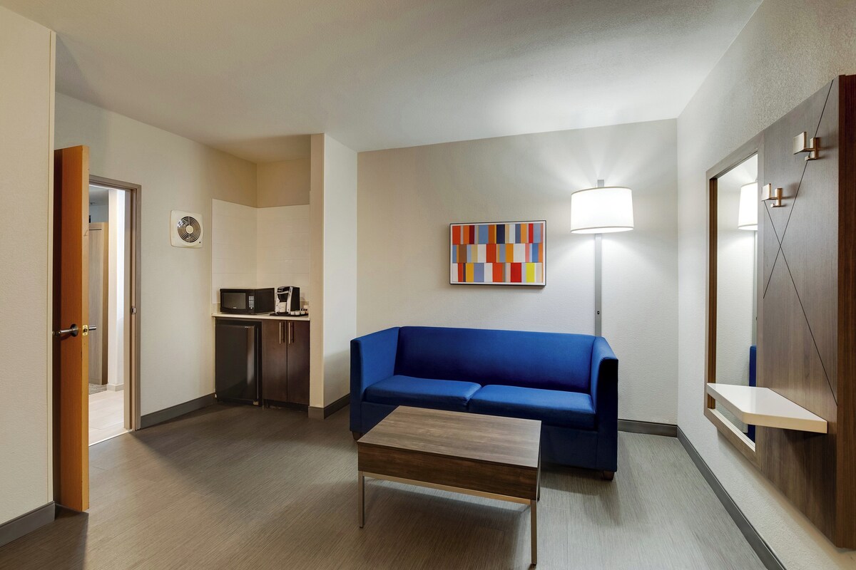 A modern hotel room with a blue sofa, a wooden coffee table, a kitchen area with a microwave, and a wall-mounted lamp.