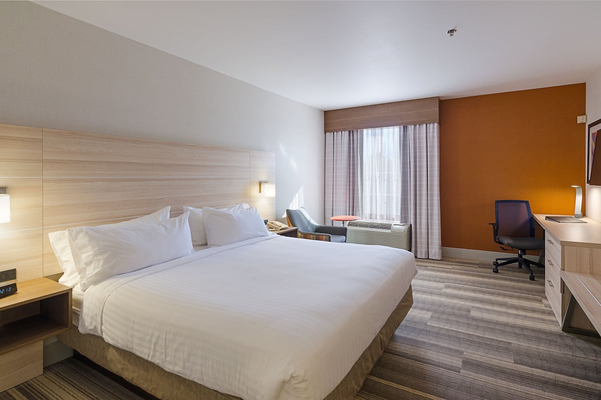 A hotel room with a large bed, a desk with a chair, a small table, and a window with curtains.