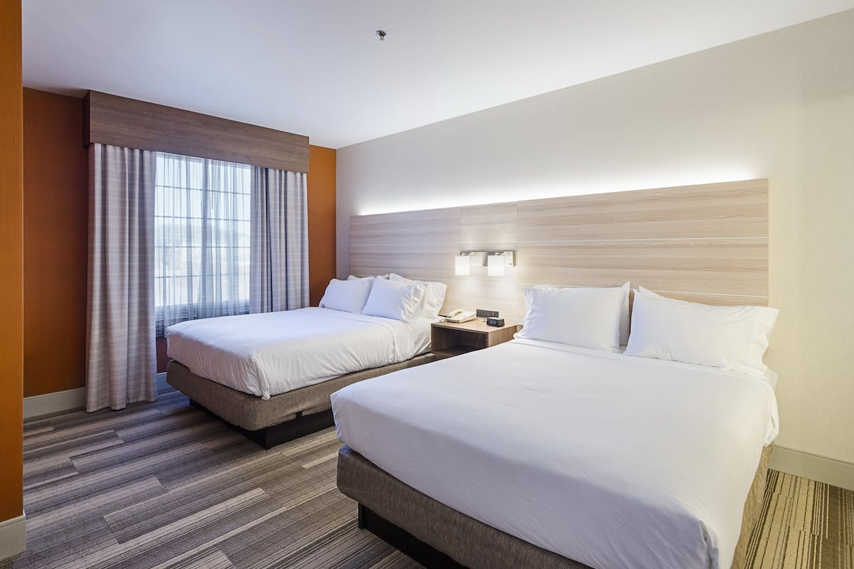 A hotel room with two beds, a large window with curtains, a bedside table with a lamp, and a wall-mounted light fixture.