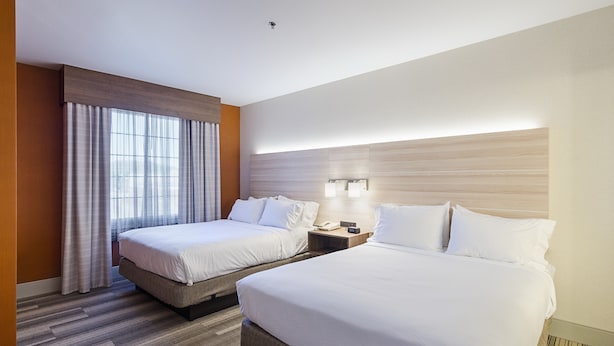 A hotel room with two beds, a large window with curtains, a bedside table with a lamp, and a wall-mounted light fixture.