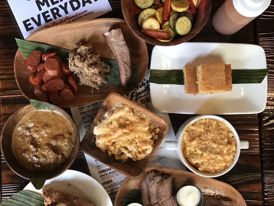 Une variété de plats, notamment des viandes tranchées, des légumes et des desserts, sont présentés sur une table en bois, avec une pancarte indiquant : « VIANDE FUMÉE TOUS LES JOURS ».