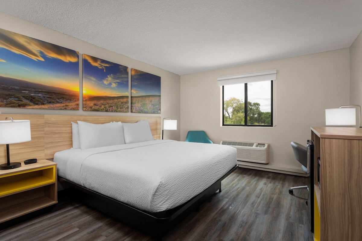 A hotel room with a bed, two bedside tables, a desk, a chair, a window, and a wall with three paintings.