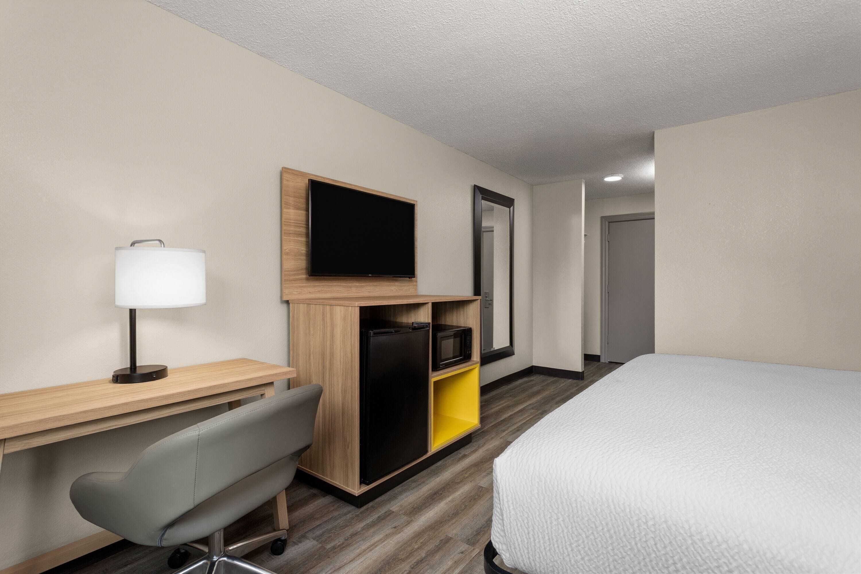 A hotel room with a bed, a desk with a lamp, a chair, a TV, and a wardrobe.