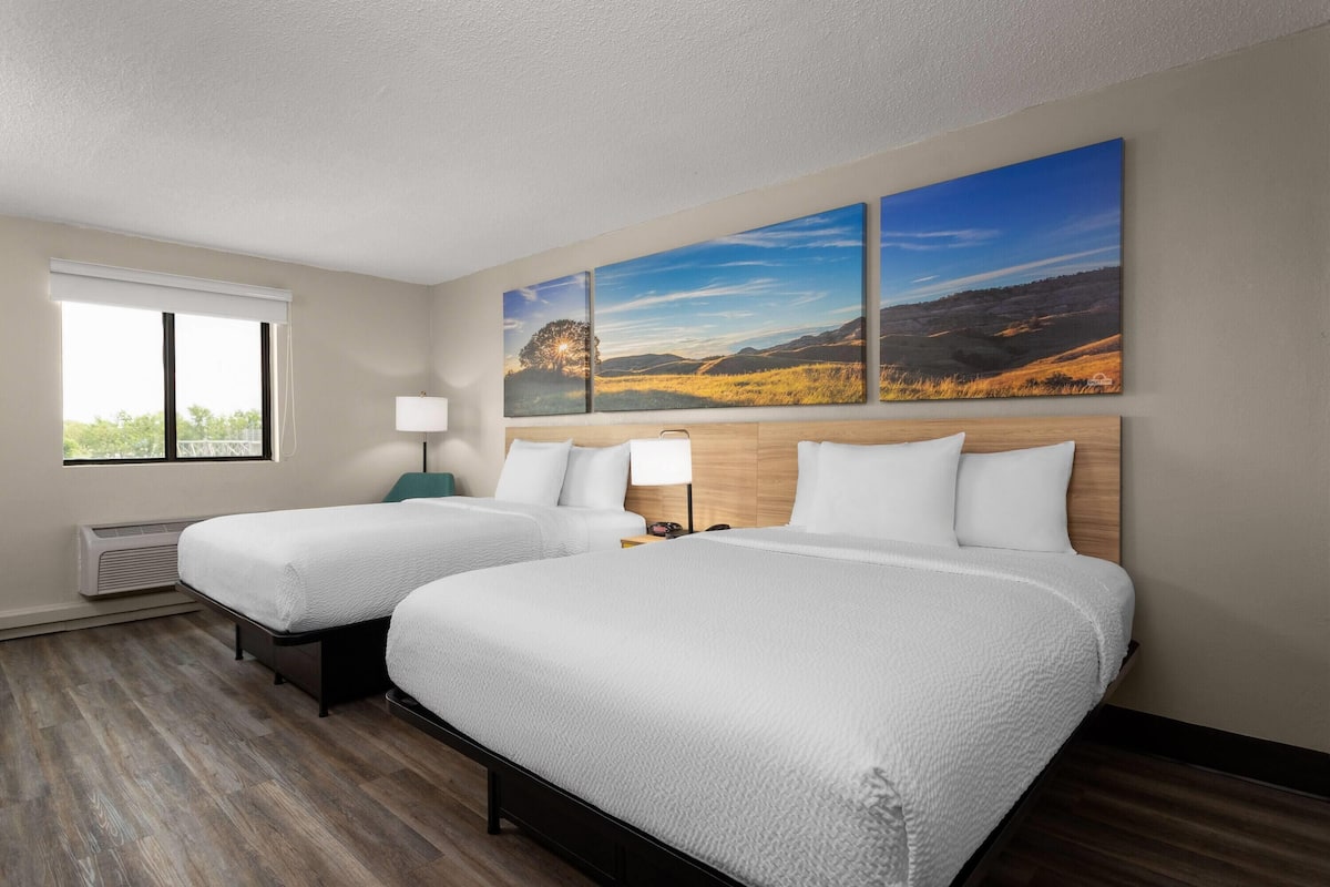 A hotel room with two beds, a large window, a wall-mounted painting, and a bedside table with a lamp.