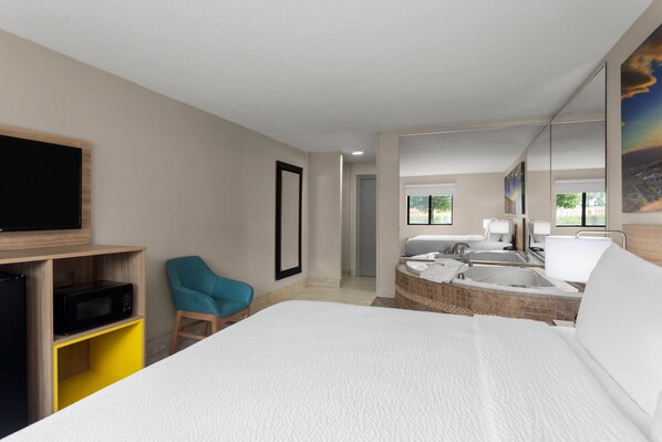 Une chambre d’hôtel moderne avec un grand lit, un téléviseur posé sur une étagère en bois, une chaise bleue, une baignoire et un grand miroir qui reflète la pièce.