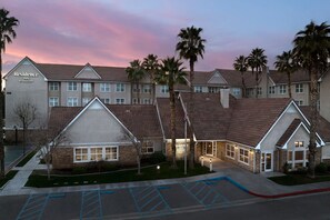 Traditional hotel with a central entrance and two wings, surrounded by palm trees and a parking lot with designated spaces.