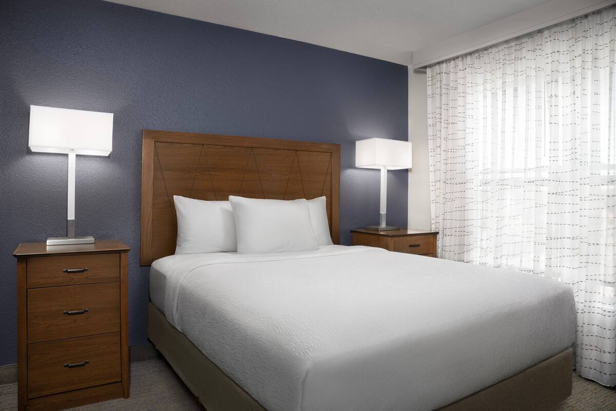 Una cama bien hecha con un cabecero de madera, dos mesitas de noche con lámparas y una ventana con cortinas de encaje.