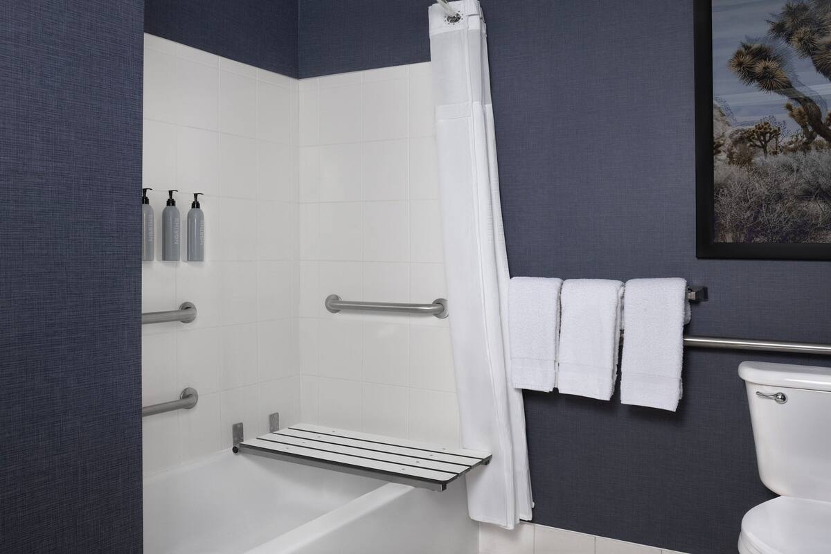 Un baño moderno con bañera, ducha e inodoro. Hay barras de apoyo, un perchero y artículos de aseo en la pared.