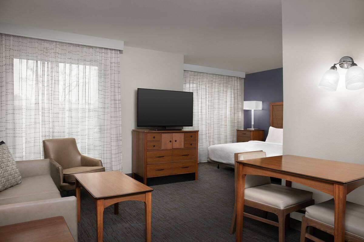 Habitación de hotel con sofá, una mesa de madera, televisión en mueble de madera, cama y mesita de noche con lámpara.