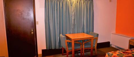 A small room with a wooden table and two chairs, a bed with a floral bedspread, a blue curtain, and an orange wall.