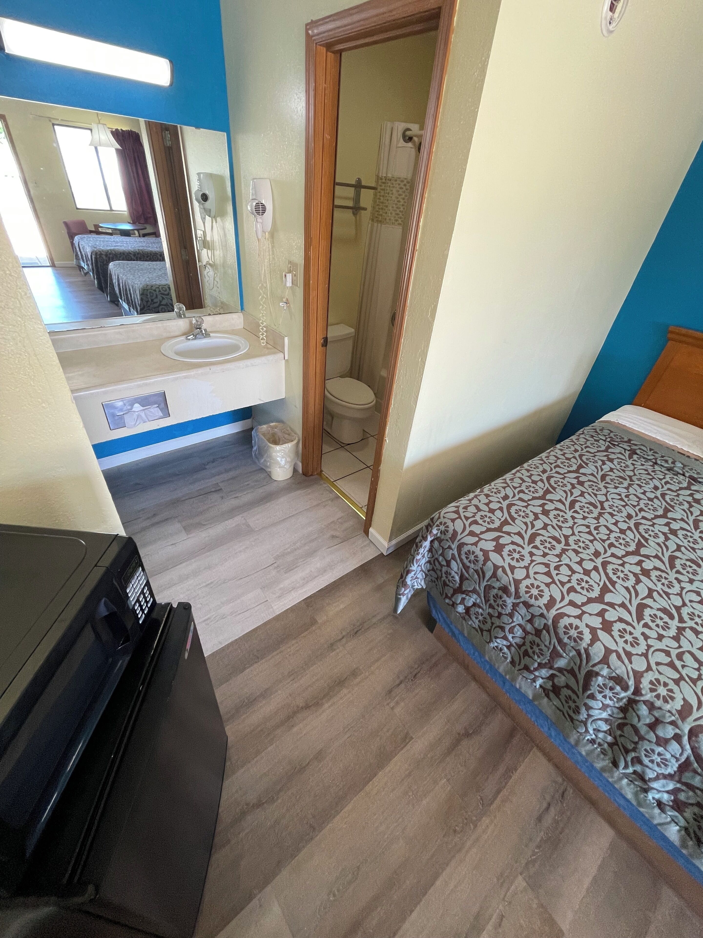 A hotel room with a bed, a desk with a computer, a bathroom with a toilet and shower, and a mirror.