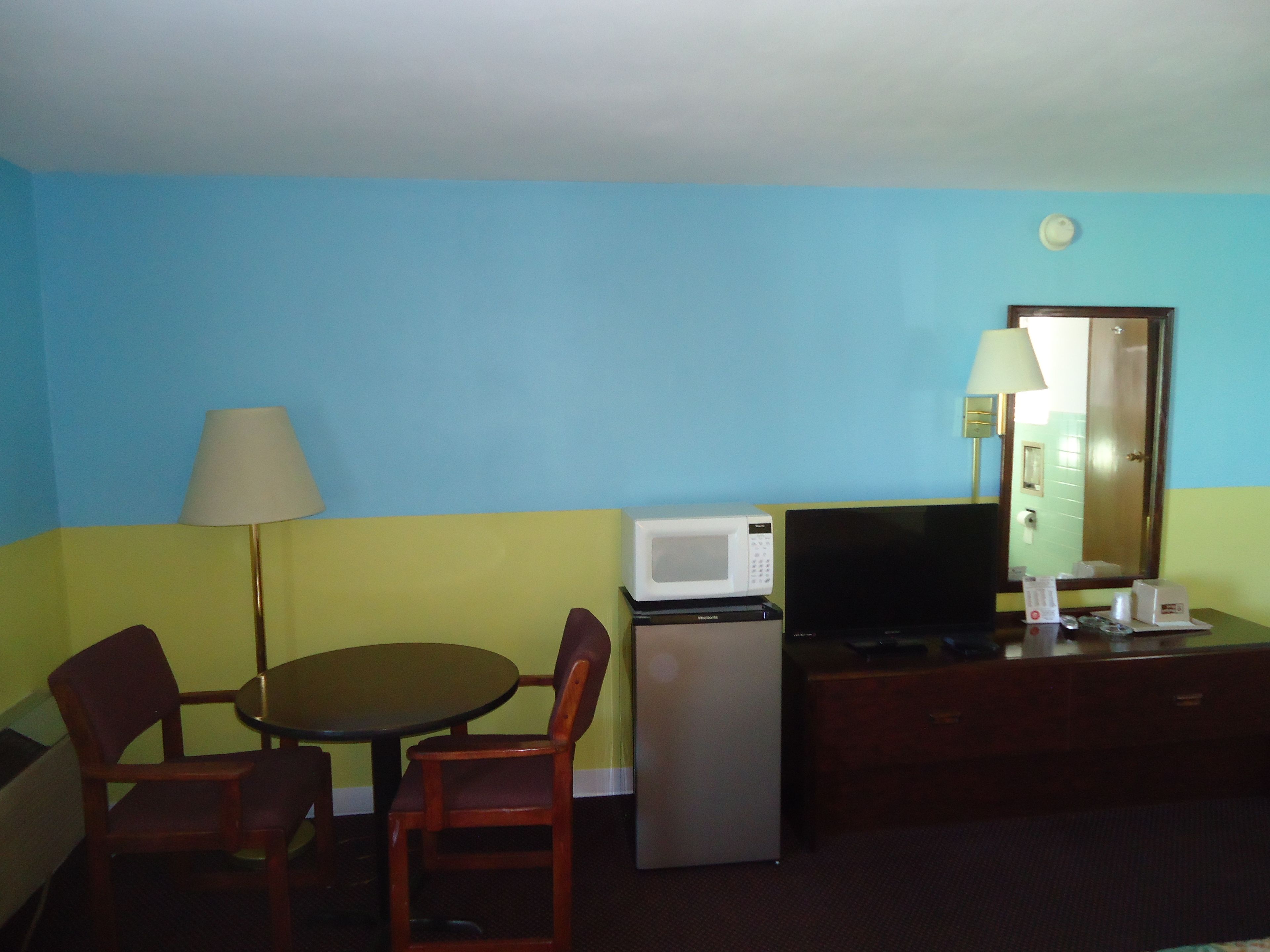 A compact hotel room with a dining table, two chairs, a small refrigerator, a microwave, a television, a lamp, and a mirror.