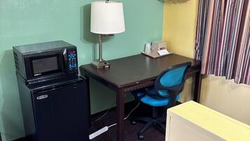 A compact hotel room with a kitchenette, a desk with a chair, a lamp, and a striped curtain.